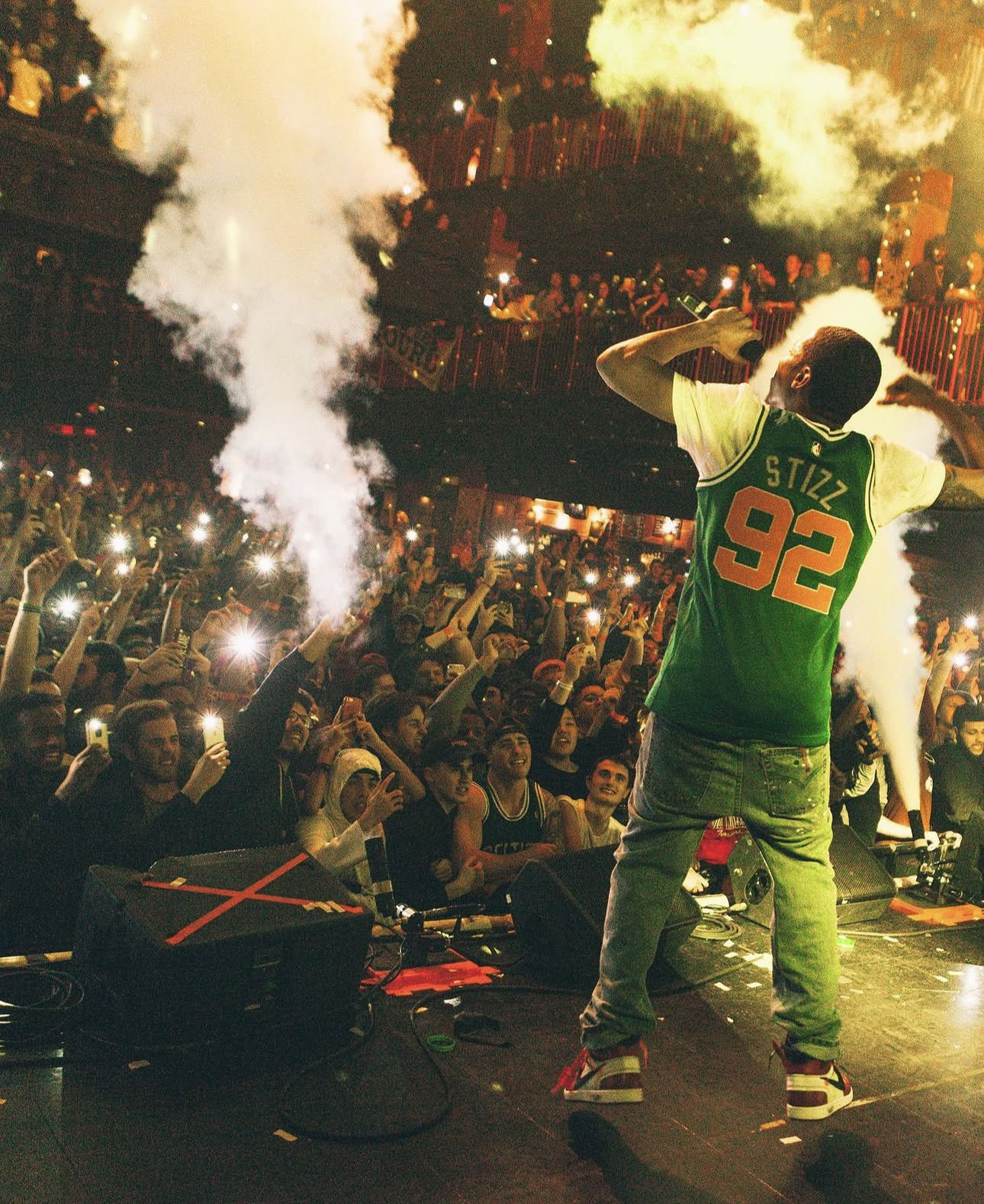 Cousin Stizz de espaldas sobre un escenario, con una camiseta verde con el nombre STIZZ y el número 92, ante un público con las luces de los teléfonos y columnas de humo