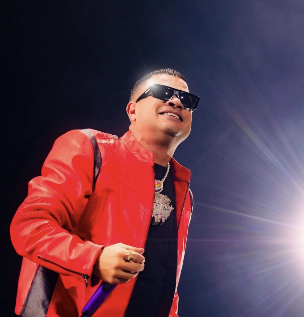 Jowell en un escenario, con chaqueta roja, lentes oscuros y micrófono en mano, bajo un destello de luz