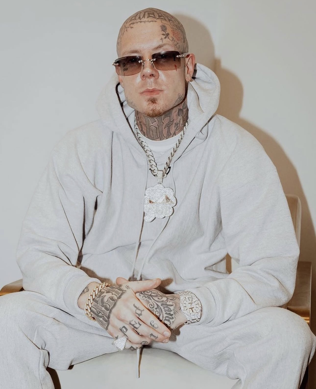 Retrato de estudio de Millyz sentado, con un conjunto gris claro, lentes de sol sin marco y una cadena de plata