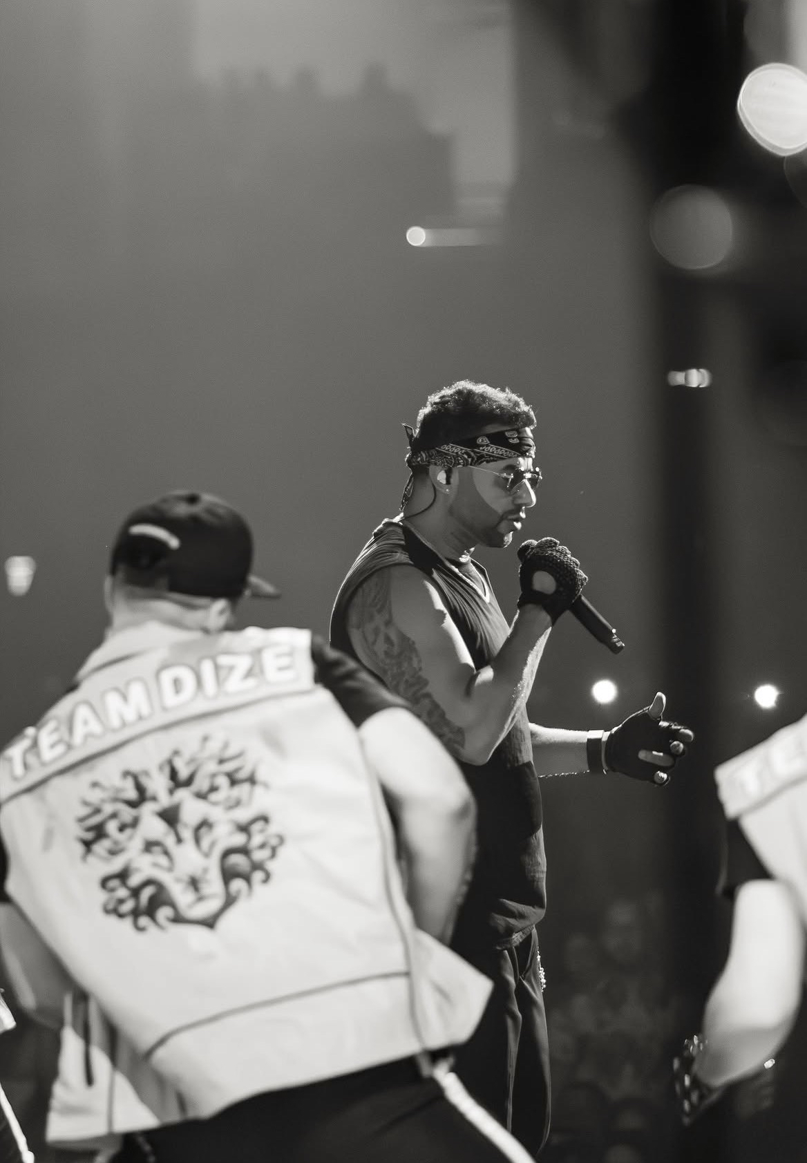 Tony Dize en escenario, en blanco y negro, con micrófono y pañuelo en la cabeza; en primer plano, un bailarín con un chaleco que dice TEAMDIZE
