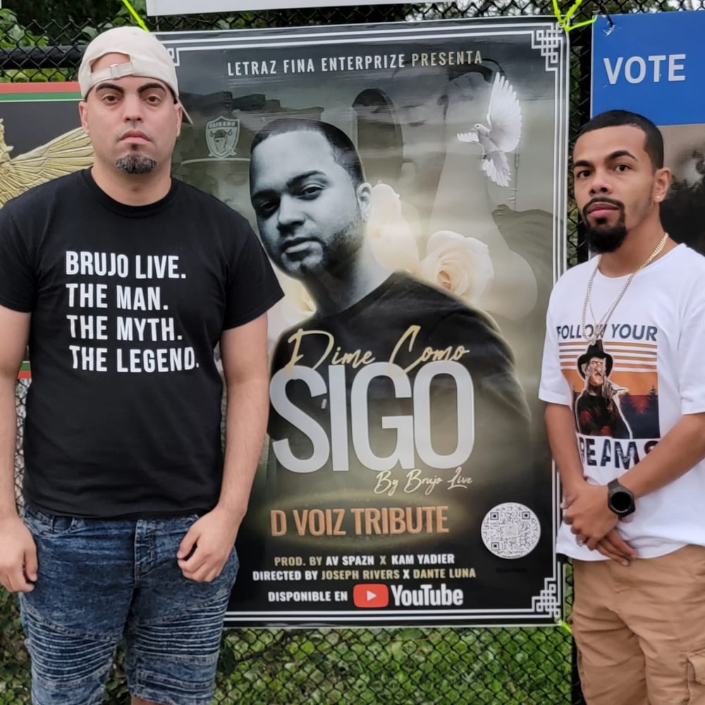 Brujo Live y Joseph Rivers junto al banner oficial del tributo a D Voiz "Dime Como Sigo." El propio banner confirma los créditos completos: producido por AV Spazn y Kam Yadier, dirigido por Joseph Rivers y Dante Luna.