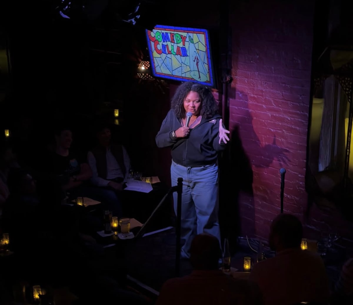 Carolina Montesquieu, con micrófono en mano, actúa en un escenario íntimo de un club de comedia con luz morada.
