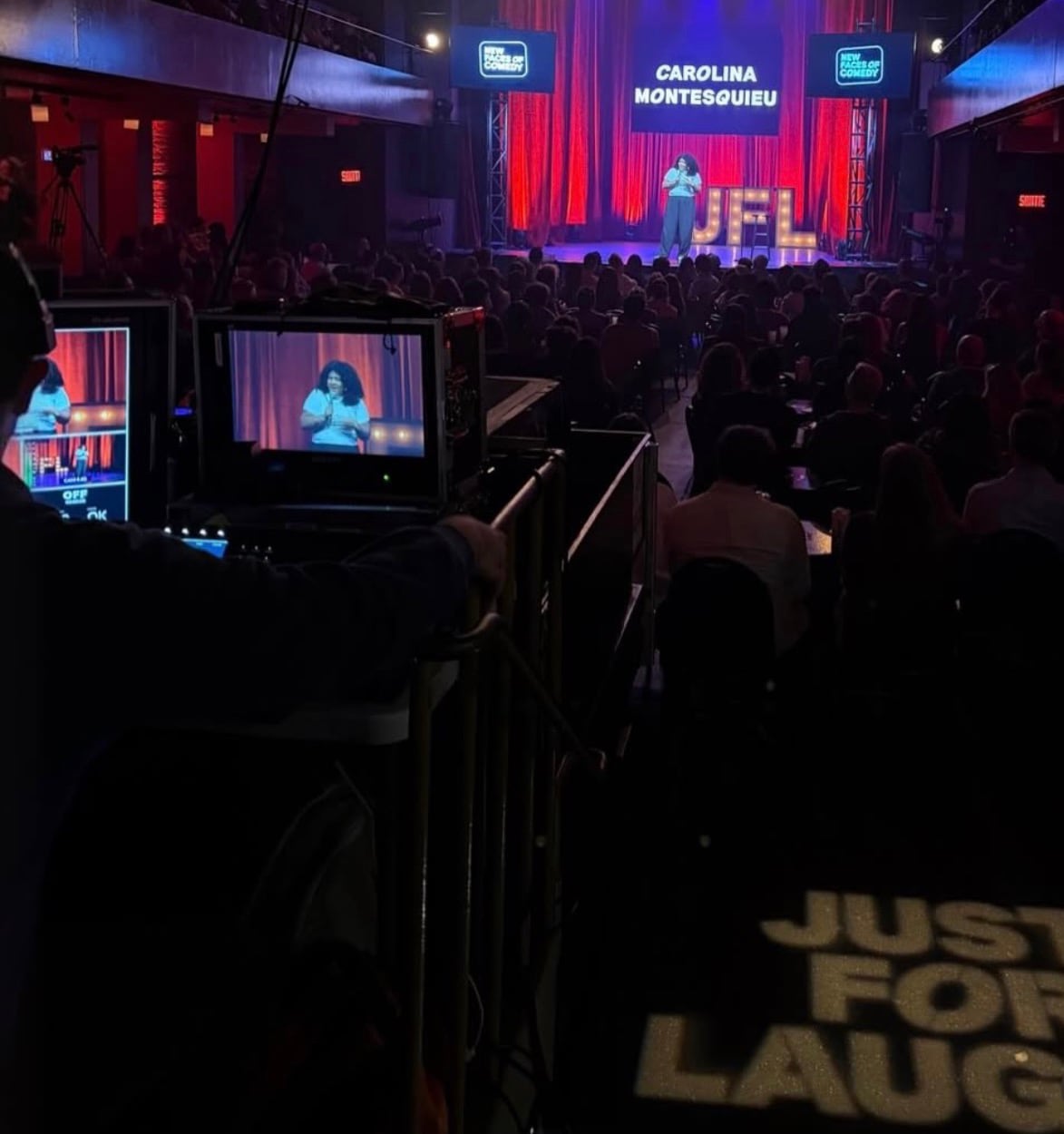 Vista desde el fondo de una sala llena: Carolina Montesquieu actúa en el escenario de New Faces of Comedy de Just for Laughs, con su nombre en la pantalla.