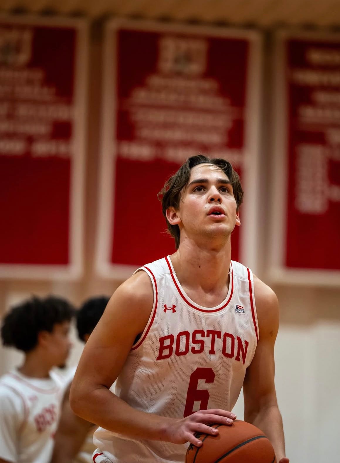 Sam Hughes tirando un libre con el uniforme de Boston University, banners de campeonato de la Patriot League detrás