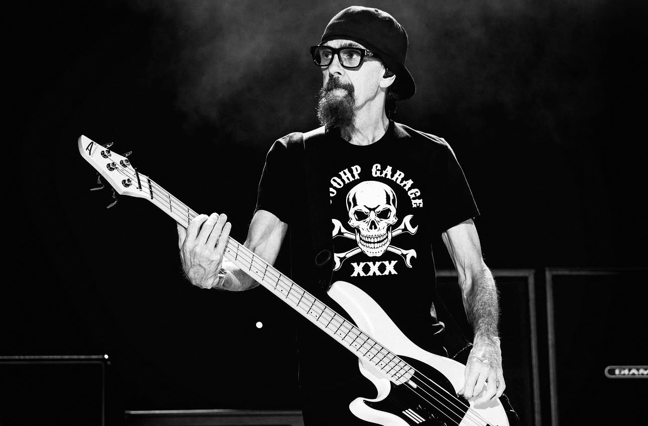 El bajista de Godsmack, retrato en blanco y negro con sombrero tipo bucket, lentes y perilla, sosteniendo un bajo blanco