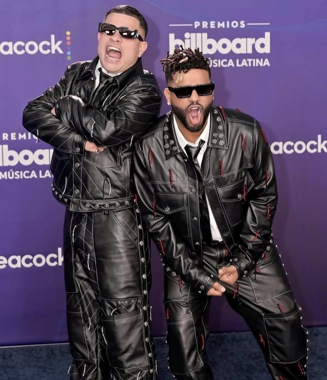 Jowell y Randy posan juntos, con trajes de cuero negro y lentes oscuros, frente al panel de los Premios Billboard de la Música Latina