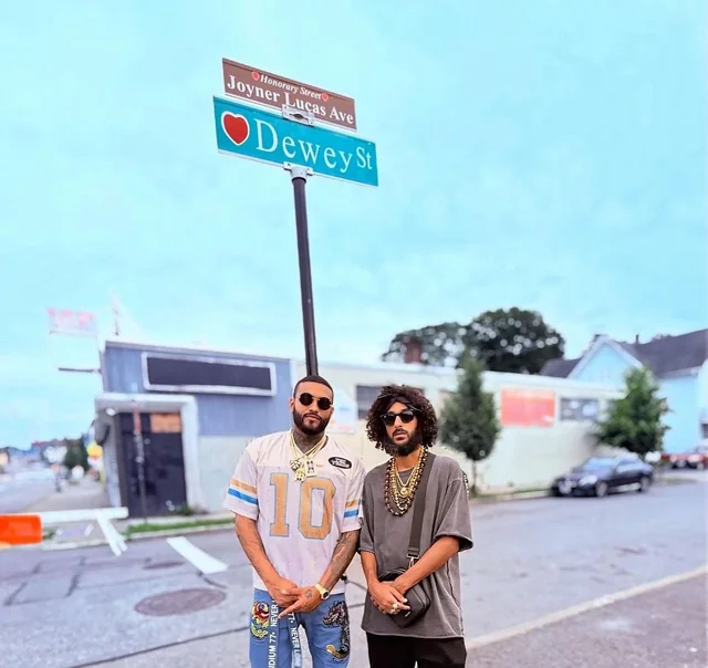 Dos hombres con lentes oscuros de pie bajo un poste con el letrero verde «Dewey St» y, encima, uno marrón que dice «Honorary Street — Joyner Lucas Ave»