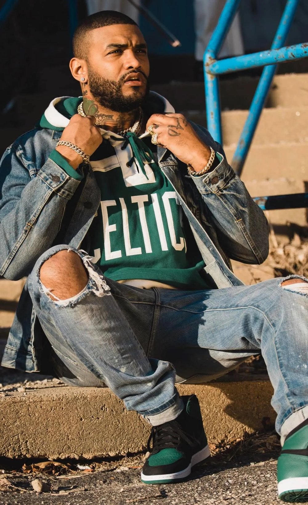 Un hombre sentado en unas escaleras de concreto, con chaqueta de mezclilla, sudadera verde con «Celtics» y jeans rotos, se sujeta el cuello de la chaqueta