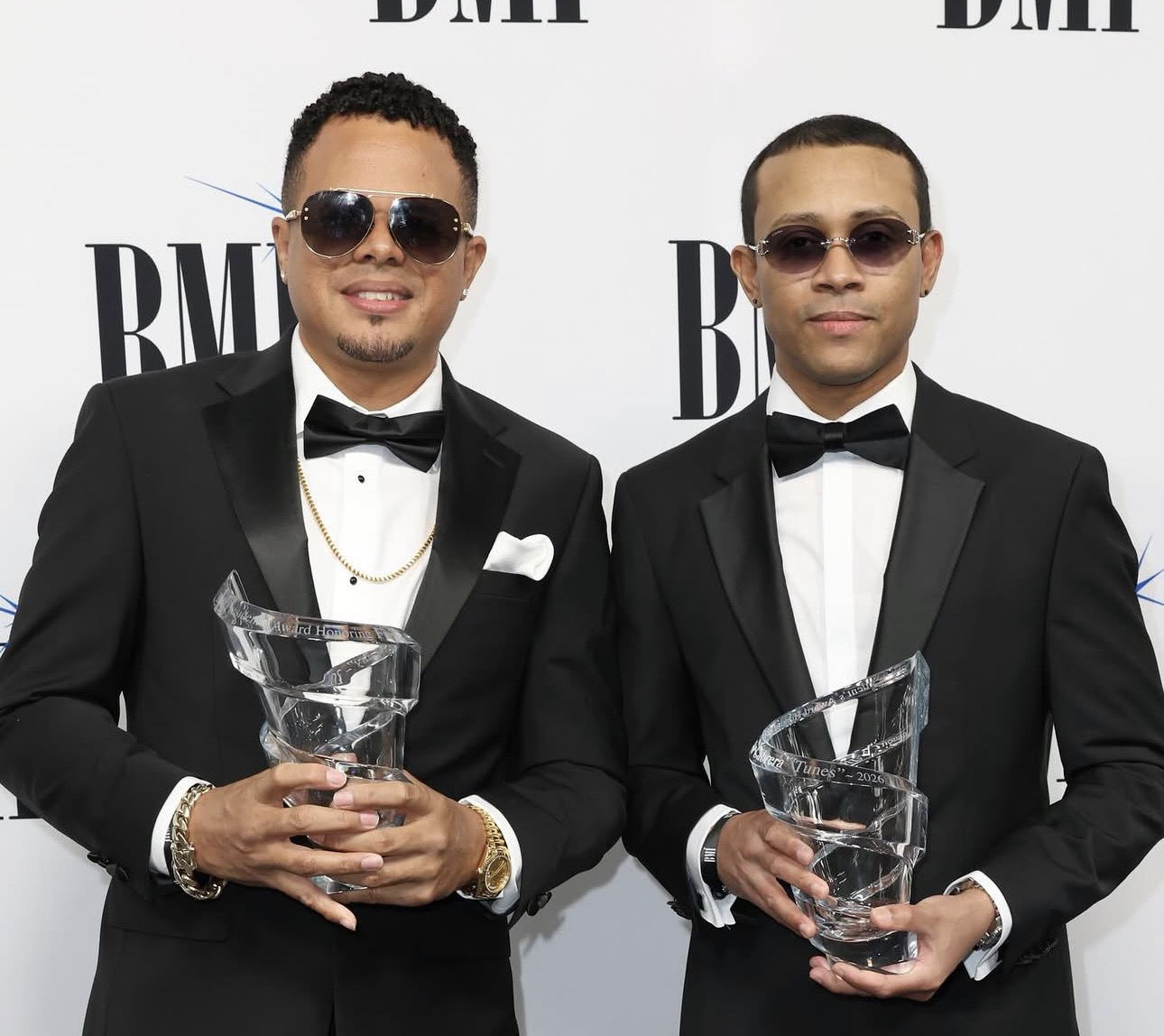 Los dos integrantes de Luny Tunes con trofeos de cristal de BMI, frente al fondo con el logo de BMI