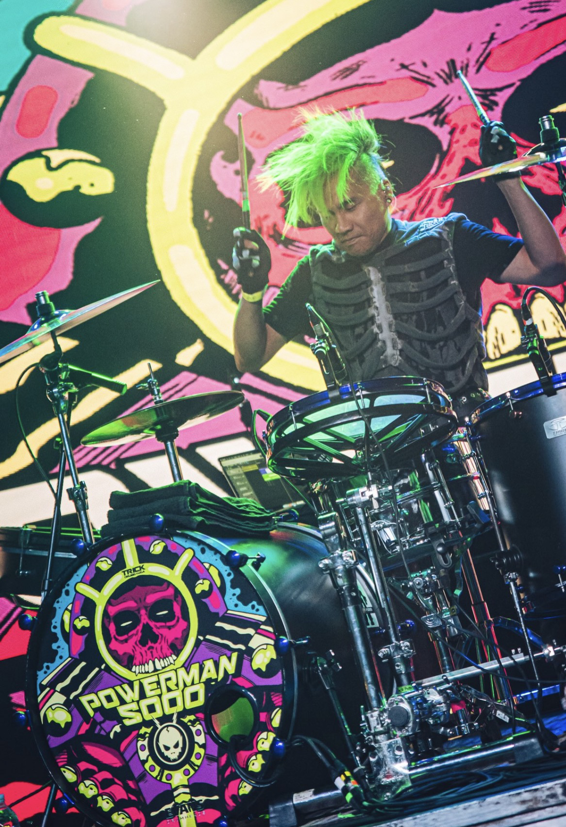 Baterista de pelo verde tras una batería cuyo bombo lleva el logotipo de Powerman 5000, ante un telón de colores estilo cómic