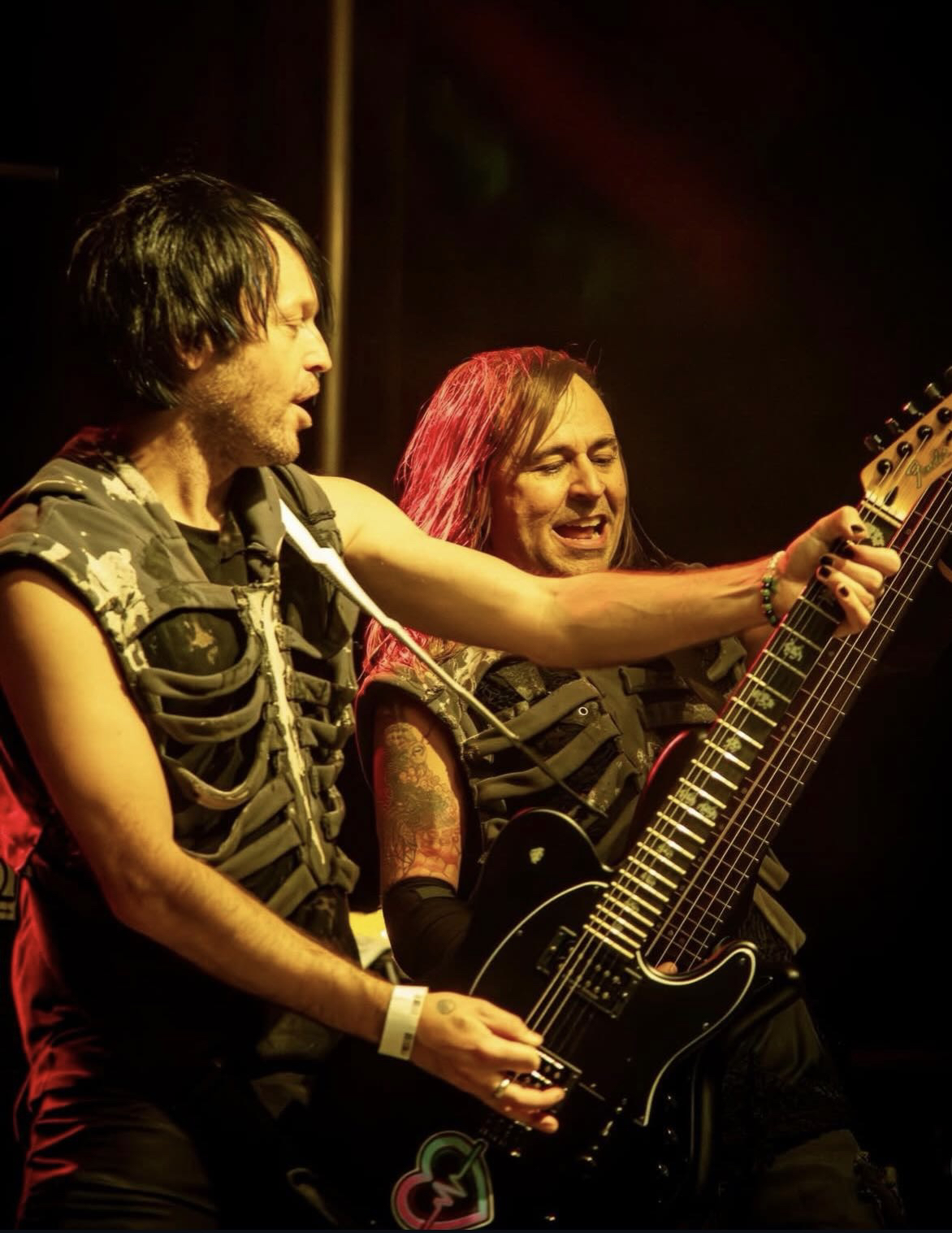 Dos músicos con chalecos de costillas, uno de ellos con guitarra eléctrica, juntos en el escenario bajo luz cálida